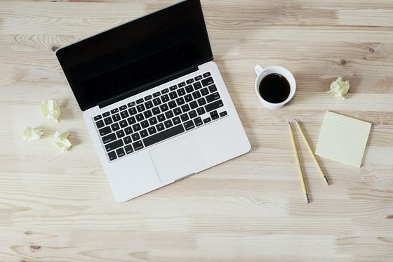 Aufgeklappter Laptop, Kaffeetasse, Bleistifte und Notizzettel auf einem hellen Holztisch