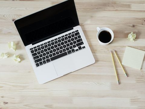 Aufgeklappter Laptop, Kaffeetasse, Bleistifte und Notizzettel auf einem hellen Holztisch