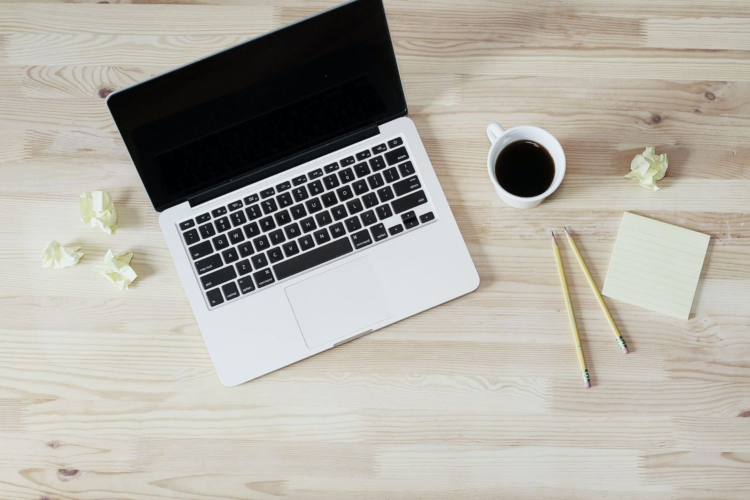 Aufgeklappter Laptop, Kaffeetasse, Bleistifte und Notizzettel auf einem hellen Holztisch