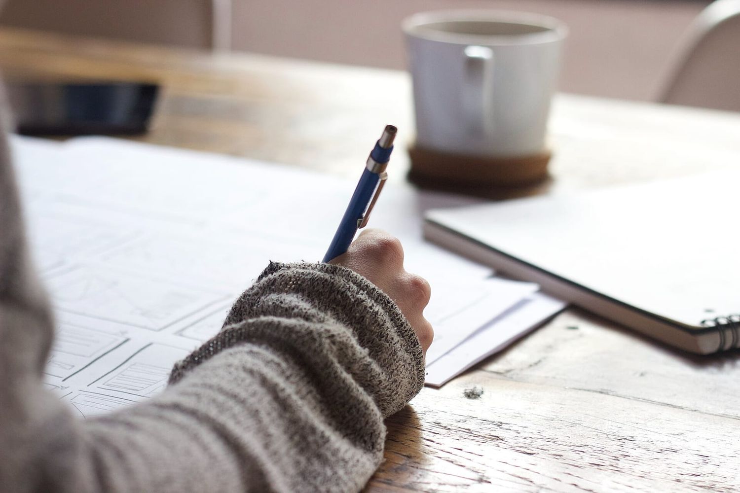 Hand mit Bleistift skizziert Seitenlayouts für eine Website, daneben Kaffeebecher und Notizbuch