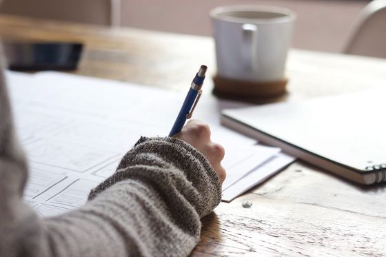 Hand mit Bleistift skizziert Seitenlayouts für eine Website, daneben Kaffeebecher und Notizbuch